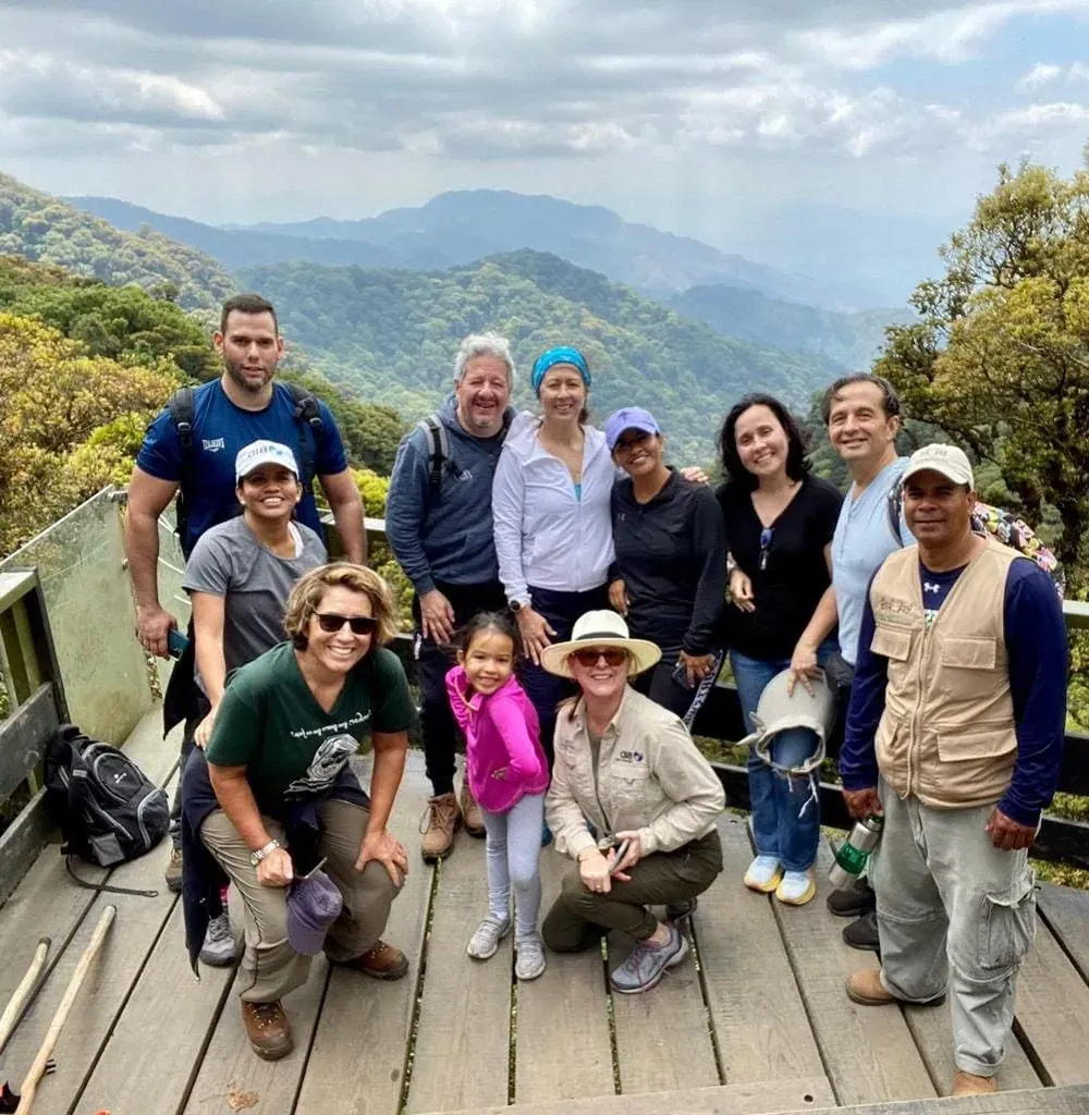 Grupo de visitantes posando en un mirador de madera con montañas en capas al fondo.
