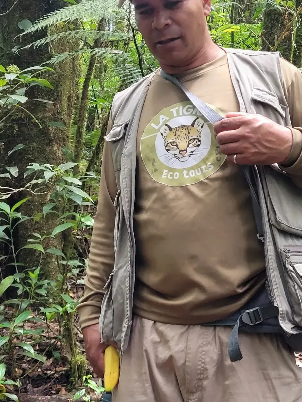 Guía con camiseta verde olivo con el logo del ocelot de La Tigra Eco Tours, de pie en el bosque.