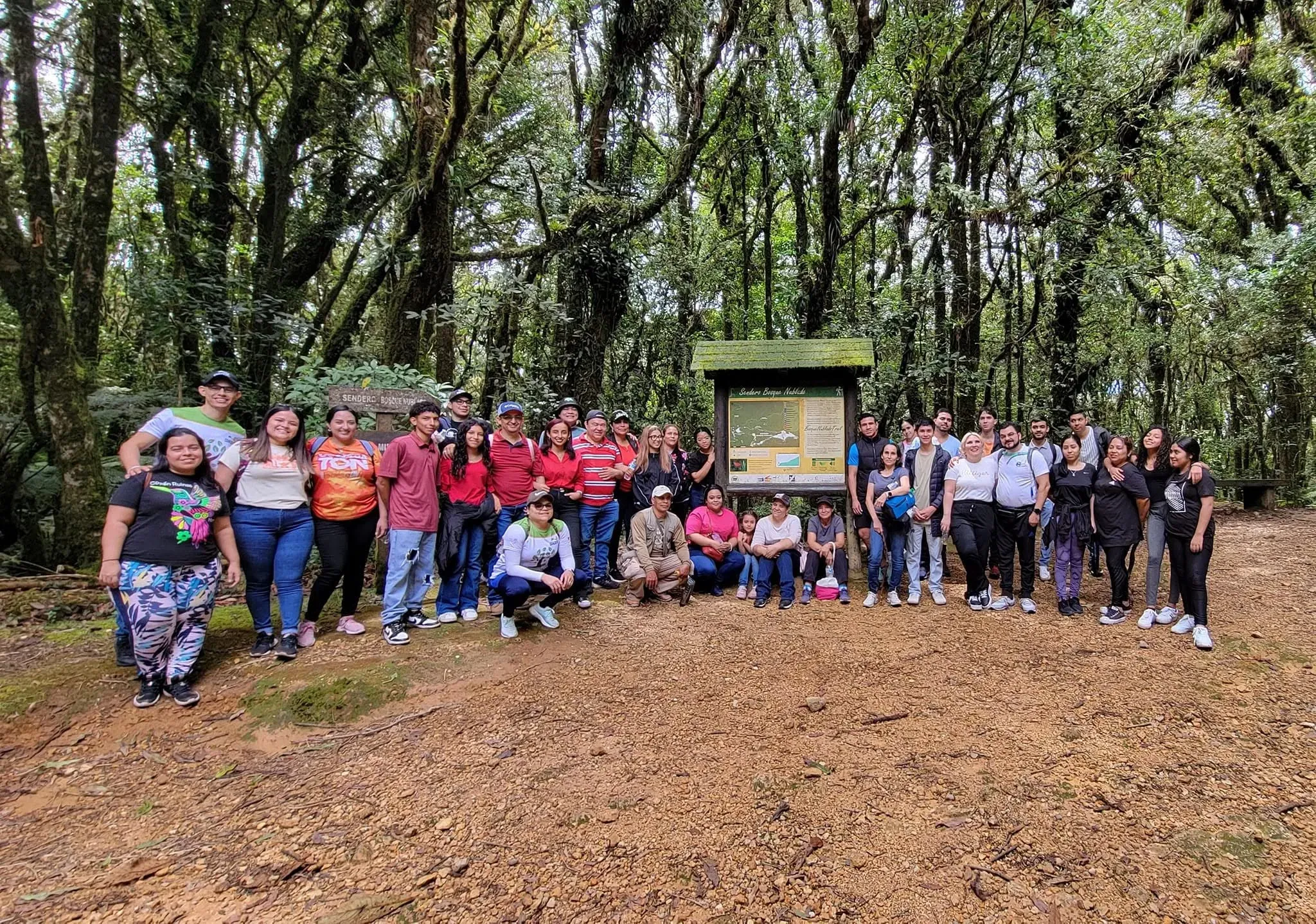 Grupo grande de visitantes posando en un claro frente al panel informativo del sendero Bosque Nublado.