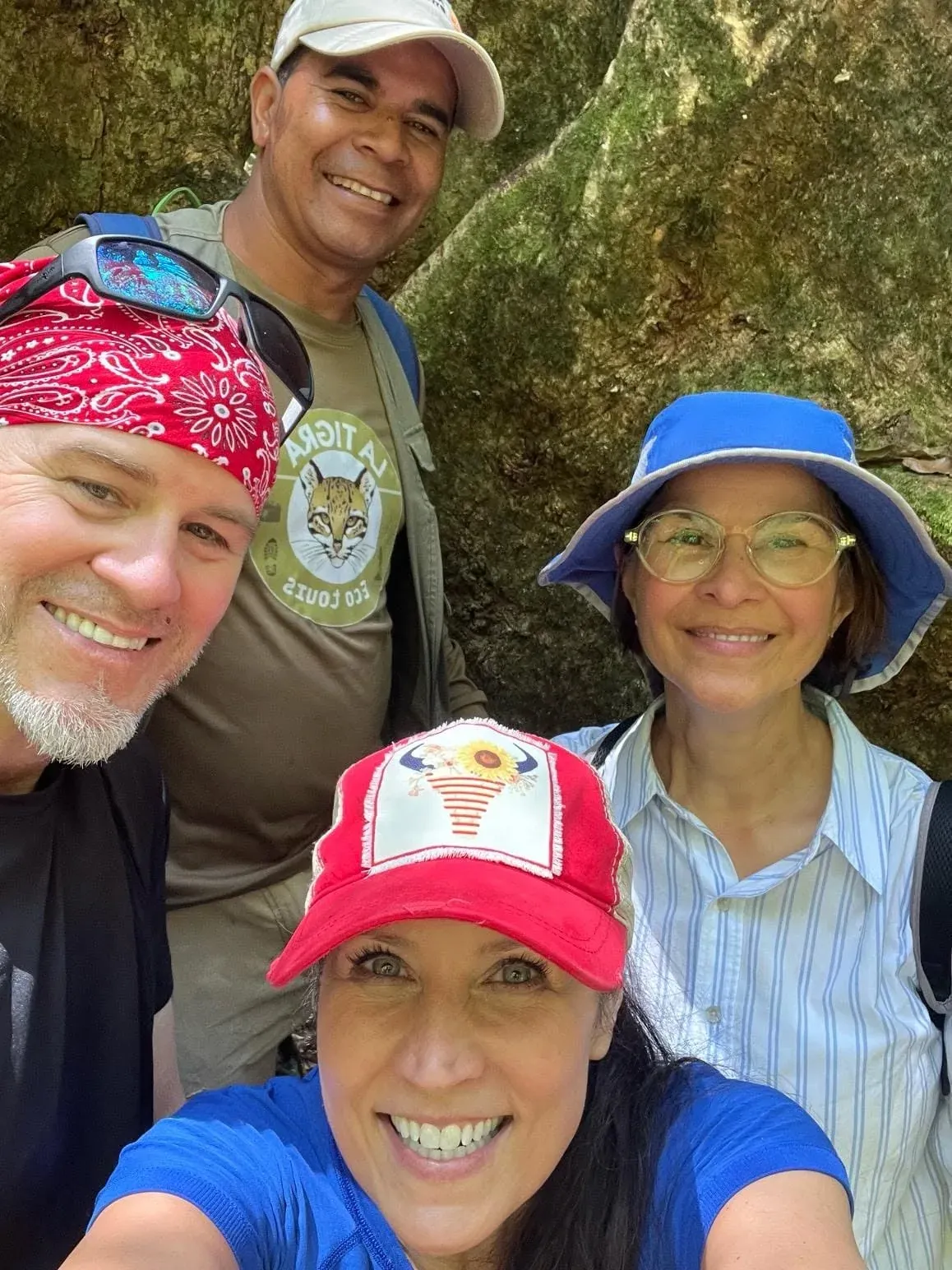 Selfie de cuatro personas sonriendo, entre ellas un guía con la camiseta del ocelot, junto a una roca cubierta de musgo.