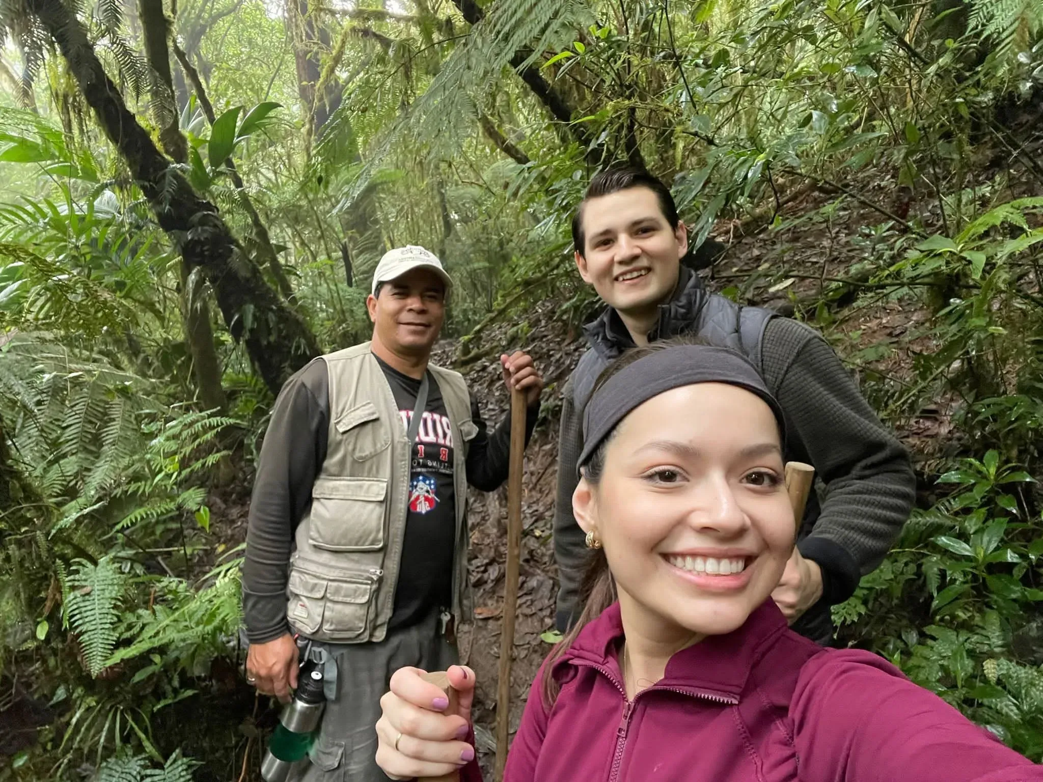 Una visitante se toma una selfie con un guía y otro visitante detrás, en un bosque de helechos.