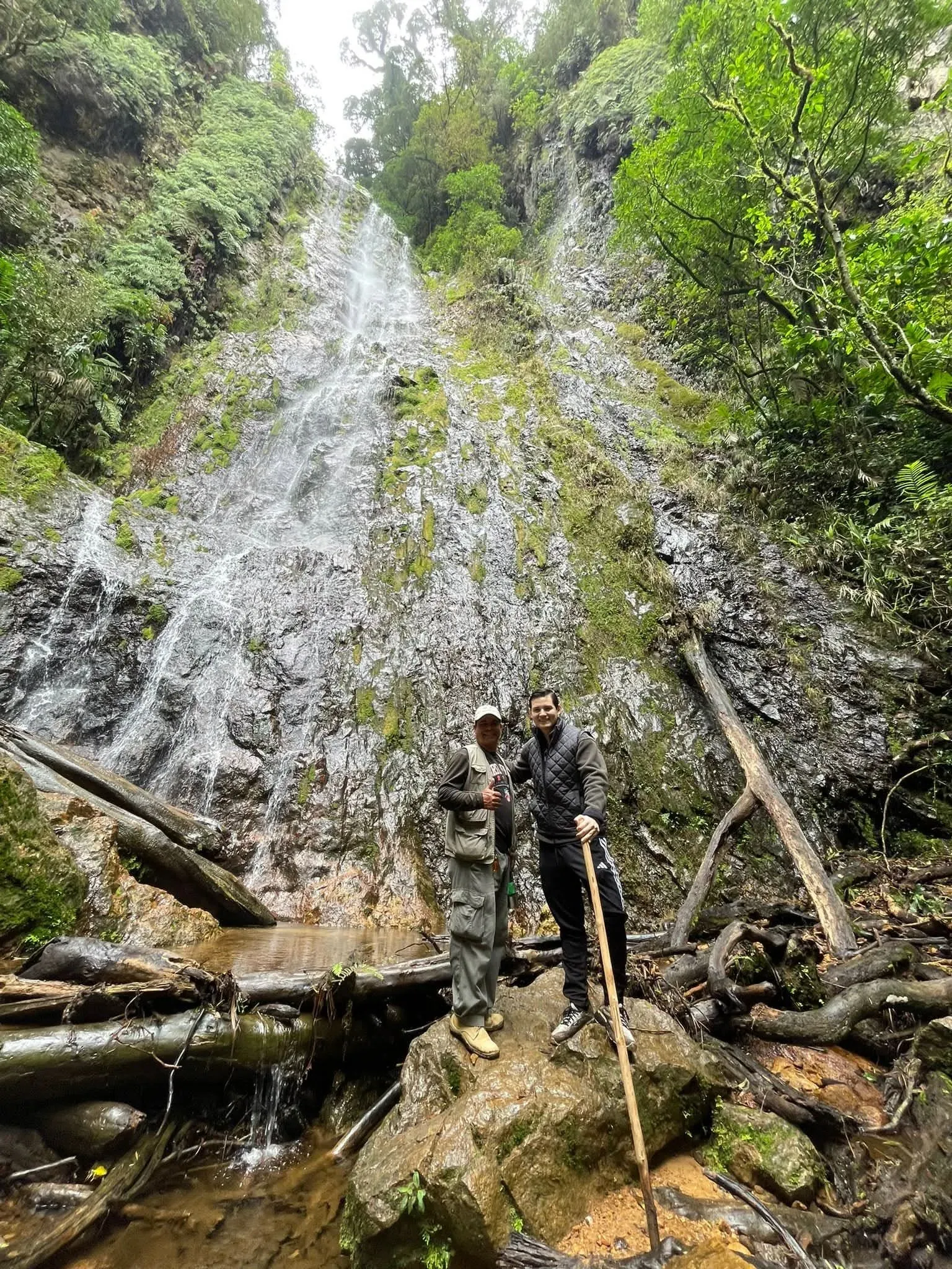Dos visitantes posan sobre una roca frente a una cascada alta que cae por una pared cubierta de musgo.