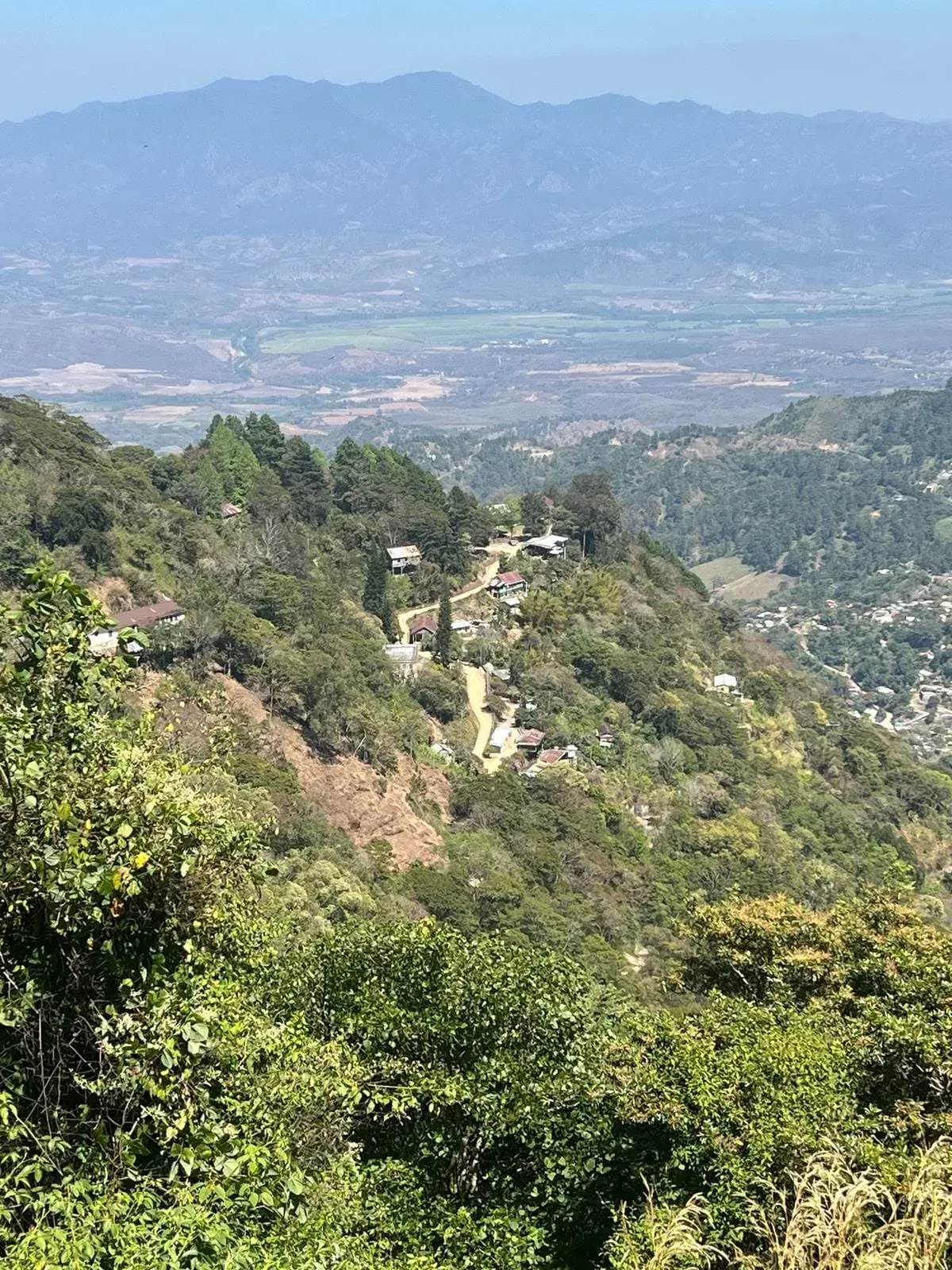 Vista desde una ladera sobre montañas boscosas, casas dispersas y un valle cultivado entre la bruma.