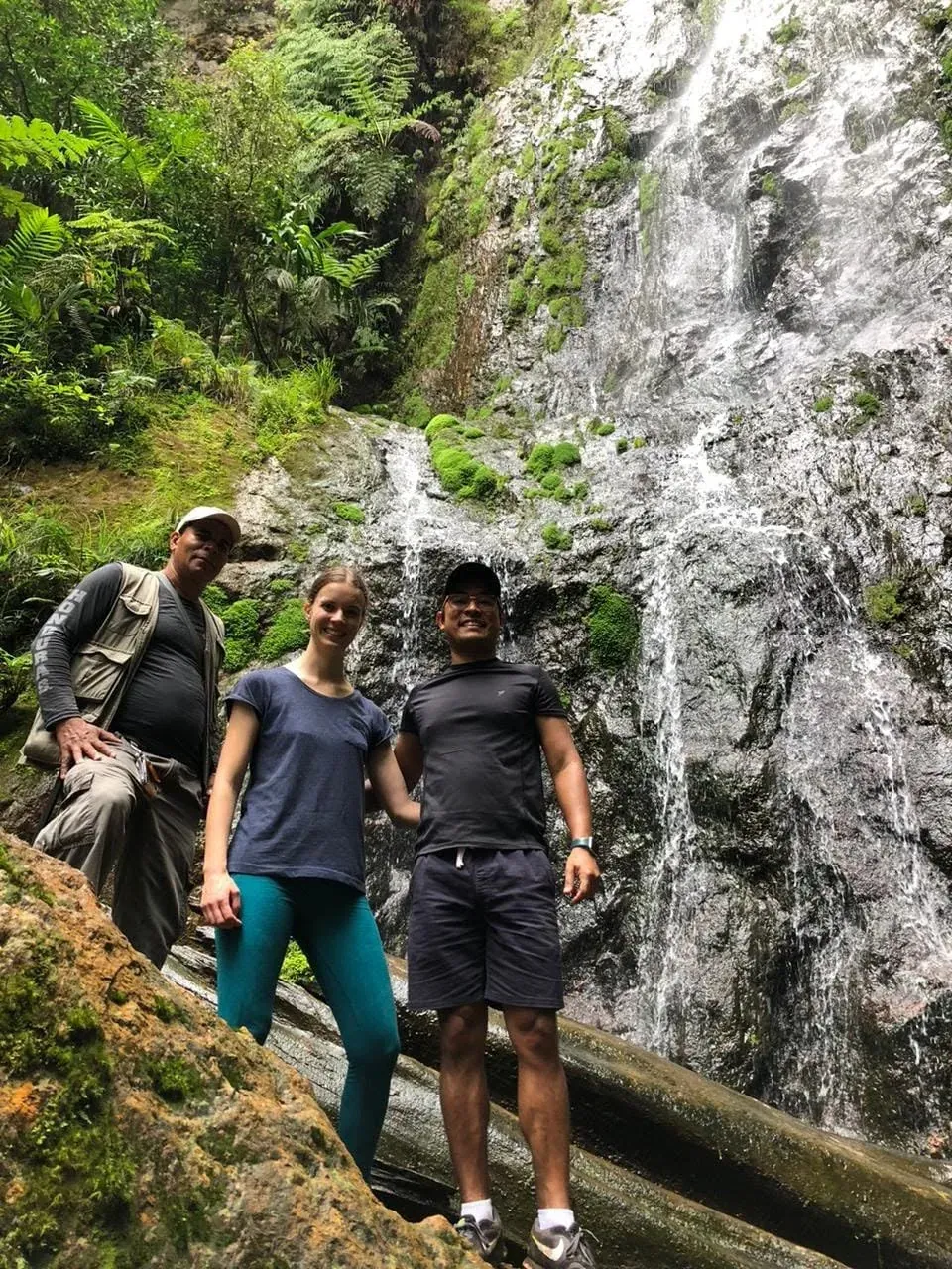 Tres excursionistas, uno de ellos guía, posan frente a una cascada alta y un tronco caído.